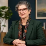 Elizabeth Fogle in a professional portrait wearing glasses and formal clothing in a modern office setting.